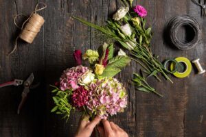 Hands arrange fresh flowers and greenery in a clear vase on a bright home table.